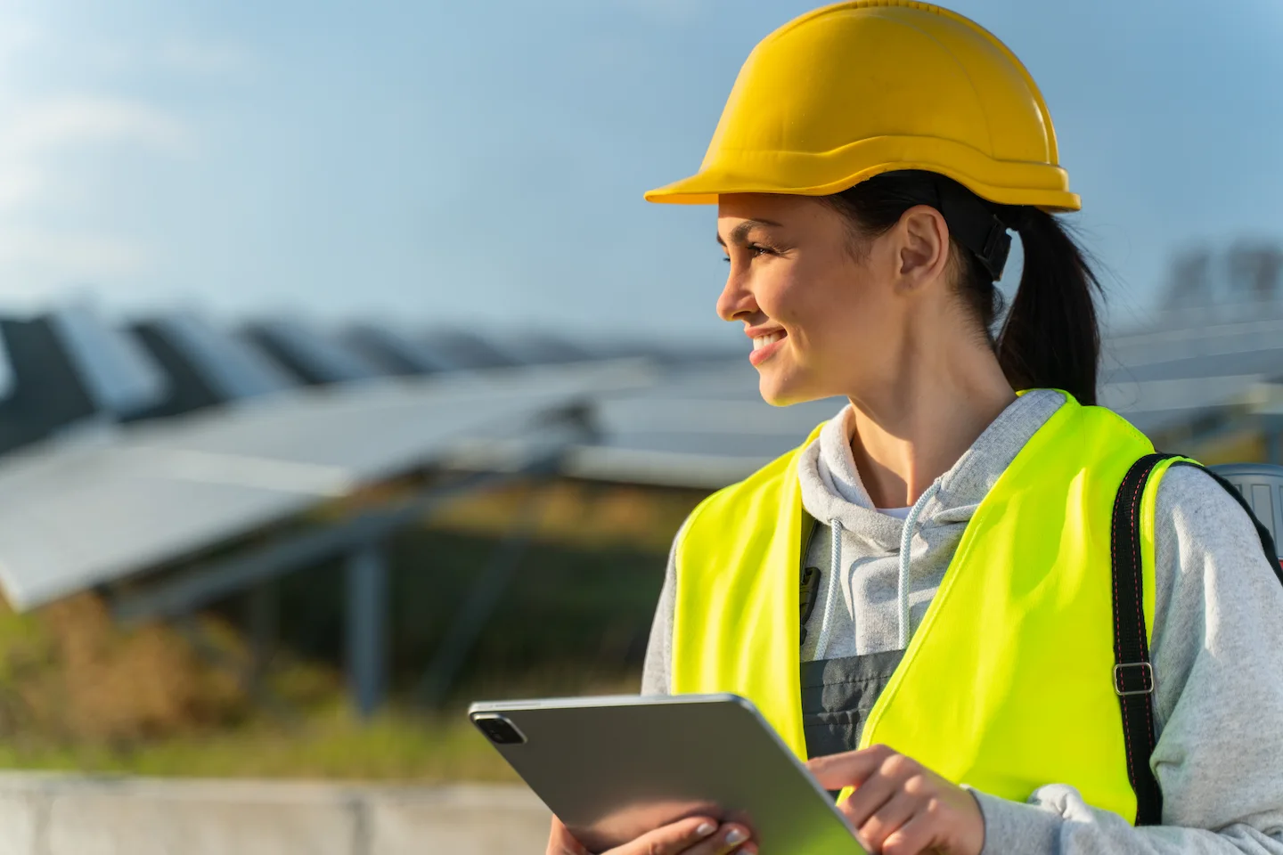 A woman in a yellow vest and hard hat smiles and holds a tablet.
